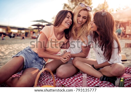 Three young happy women friends in casual dressed speaking with each other on sandy beach on sunny day.