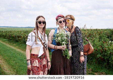 Three young hippie women, wearing boho style clothes, standing on green currant field, holding camomiles' bouquet in summer. Eco tourism concept. Friends traveling in rural countryside.