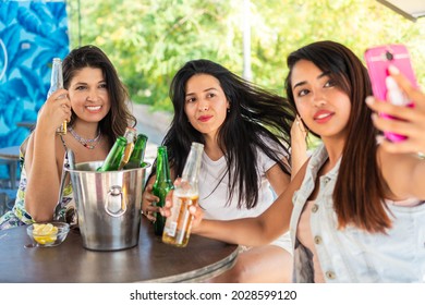 Three young latin friends having their picture taken while drinking beers. Girl friends enjoying a nice summer evening on a terrace. Girl friends taking a selfie for their social networks.