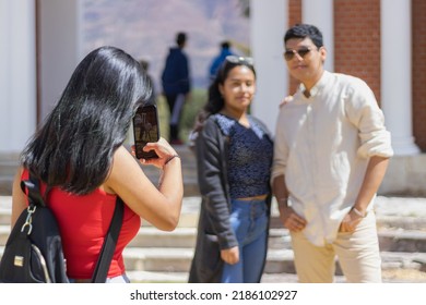 Three Young Latino Friends Enjoying Their Vacation, A Woman Taking A Picture With Her Cell Phone Of Her Two Friends At Noon At The Back Of The Church In Campo Santo. Two Friends Posing For A Photo.