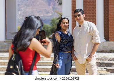 Three Young Latino Friends Enjoying Their Vacation, A Woman Taking A Picture With Her Cell Phone Of Her Two Friends At Noon At The Back Of The Church In Campo Santo. Two Friends Posing For A Photo.