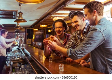 Three young men in casual clothes are talking, laughing and clanging glasses of alcoholic beverage together while sitting at bar counter in pub