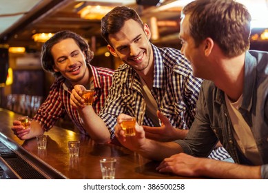 Three Young Men In Casual Clothes Are Talking, Laughing And Drinking While Sitting At Bar Counter In Pub