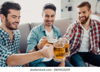Three young men sit on a couch, smiling and toasting with cold beer mugs during a relaxed at-home gathering that conveys friendship, celebration, leisure and good times.