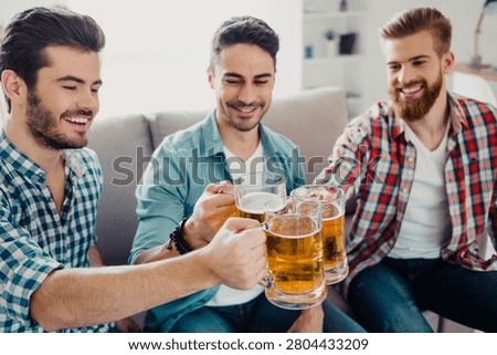 Three young men sit on a couch, smiling and toasting with cold beer mugs during a relaxed at-home gathering that conveys friendship, celebration, leisure and good times.