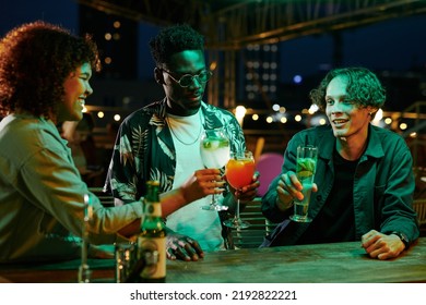 Three Young Multicultural Friends In Casualwear Toasting With Cocktails Over Bar Counter And Enjoying Night Party In Rooftop Cafe