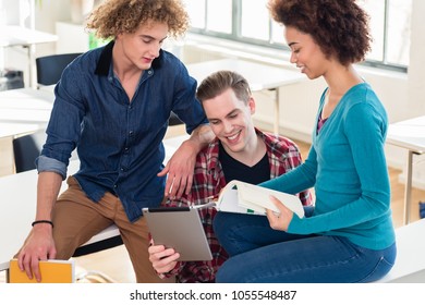 Three young students using both a paper book and a tablet PC for checking information during break in the classroom of a modern college