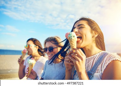 Three young woman eating ice cream cones at the seaside as they stroll along a waterfront promenade on a hot summer day during their vacation