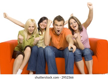 Three Young Woman And A Man Are Sitting On The Orange Couch And Looks Like Watching TV. Man Is Holding Remote Control. The're Very Excited By What They're See. Isolated On White In Studio.