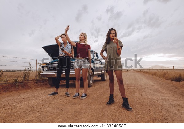 Three young woman stranded in middle of a\
countryside road with broken down car using their phones to call\
emergency assistance.
