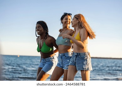 Three young women in bikinis and denim shorts laughing and walking along the beach, enjoying a sunny day. - Powered by Shutterstock