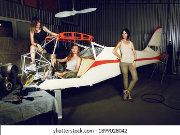 Three Young Women With A Body Plane In Airplane Hangar Near The Table With Engine And Tools. Beautiful Female Workers In Small Aviation. Advertising Image Of Aircraft Making Or Maintenance.