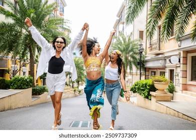 Three young women of diverse backgrounds joyfully holding hands and running down a sunny, palm-lined street in a vibrant, urban neighborhood. Their laughter and energy capture the spirit of friendship - Powered by Shutterstock