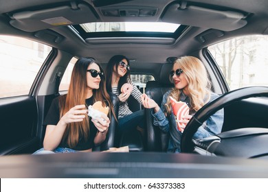 Three Young Women Drive A Car, Speaking Each Other And Eating A Fast Food In A Car In Traffic