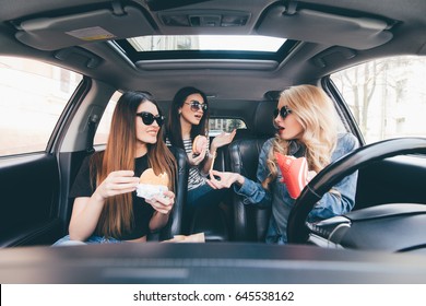 Three Young Women Drive A Car, Speaking Each Other And Eating A Fast Food In A Car In Traffic