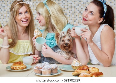 Three young women gossipers and little dog sit at table with baked sweets in kitchen.