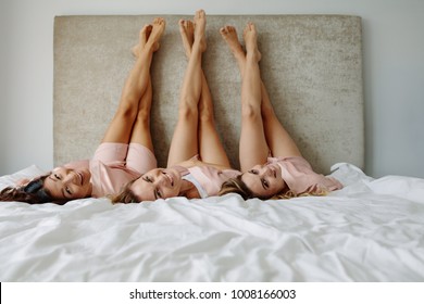 Three Young Women Lying On The Bed With Legs Up. Women In Sleeping Robe Lying In Bedroom With Legs Raised Up.