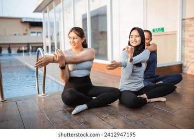 three young women sitting crosslegged doing arm stretches near a hotel swimming pool
