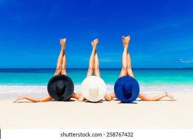 Three Young Women In Straw Hats Lying On A Tropical Beach, Stretching Up Slender Legs. Blue Sea In The Background. Summer Vacation Concept.