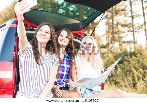 Three\
young women with suitcases on a trip by car. They sit in the back\
of the car, they look at the map, resting after a long drive and\
having fun. Hitchhiking and car trips with\
friends