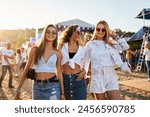 Three young women walk together on sunny beach at music festival. Happy friends in trendy summer outfits enjoy live concert, dancing, laughing in festive atmosphere. Group of girls having fun by sea.