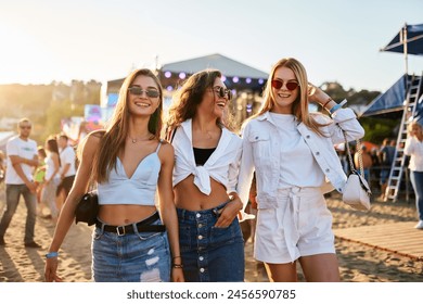 Three young women walk together on sunny beach at music festival. Happy friends in trendy summer outfits enjoy live concert, dancing, laughing in festive atmosphere. Group of girls having fun by sea. - Powered by Shutterstock