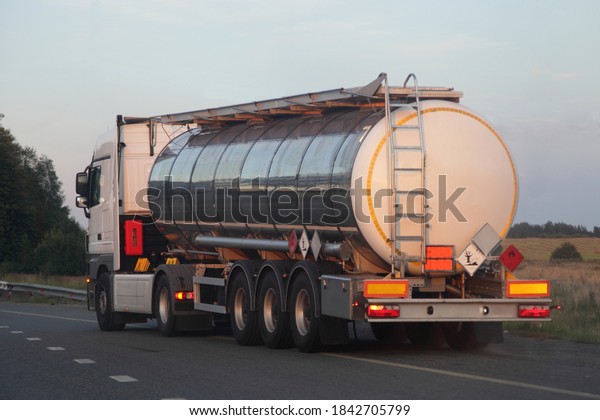 Three-axle semi truck with shiny chrome barrel tank\
with clear place on ADR sign moving on empty suburban asphalt\
highway road on a summer day on blue sky, dangerous cargo\
logistics, back side\
view