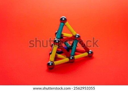 A three-dimensional triangle structure crafted from vibrant magnetic building sticks and spheres rests on a bright orange background. The design showcases creativity and engineering principles.