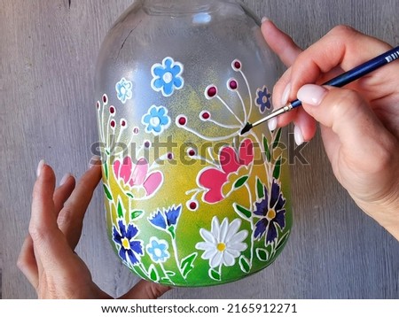A three-liter jar with stained glass painting. A frame from the video of the drawing master class. Artist's hands with a brush. The process of applying paint. Home hobby needlework of women housewives