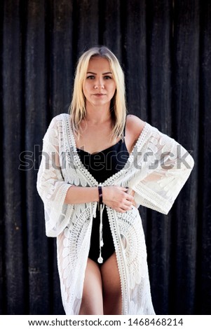 Three-quarter portrait of blond woman, wearing ivory lace peignoir and black swimsuit at vacation in bungalow hotel. Young attractive girl, posing in front of black wooden columns in tropical resort