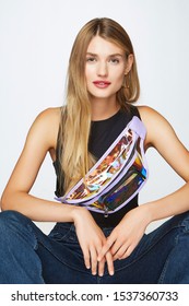 Three-quarter Shot Of A Blond Girl, Wearing Black Tank Top And Blue Jeans. She Has Clear Holographic Belt Bag With Pocket On The Front Side Over Her Shoulder And Big Hoop Earrings. 