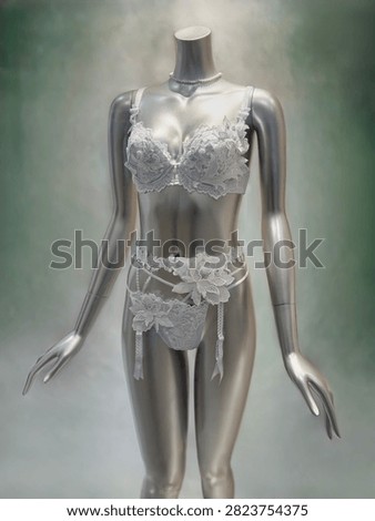 A three-quarter vertical shot featuring a metallic silver mannequin presenting an ornate white lace lingerie set. The ensemble includes a structured underwire bra, matching lace panties, and a decorative garter belt styled with large 3D floral appliqué accents and dangling ribbons. Complemented by a pearl necklace against a soft, muted greenish-grey backdrop, this display highlights delicate embroidery and luxury craftsmanship. Ideal for stock media on women's intimate apparel, luxury lingerie, bridal underwear, boutique fashion displays, and romantic clothing themes.