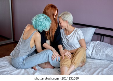 Threesome Concept. Three Lgbt Caucasian Ladies In Domestic Clothes Sit On Bed Flirting, Hugging, Women In Love, Lesbian, Same Sex Females Enjoy Spending Time Together, Going To Have Sex. Side View