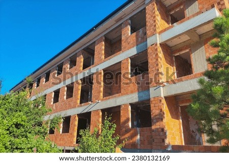 Three-storey building under construction made of red brick and concrete with a row of growing trees. Concept of construction, growth that does not harm the environment, flora and fauna. Ecofriendly.