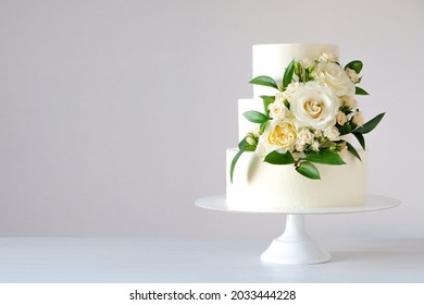 Torta de boda blanca de tres niveles decorada con flores y hojas verdes sobre un fondo de madera blanca.