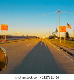 Three-wheel Motorcycle Riding In Texas Highway At Sunset. Evening Landscape With Long Shadow Of Motorbike On Asphalt, Road Signs And American Flag.  