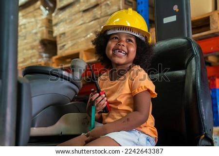 three-year-old African-American girl in an engineer's helmet smiling happily drives a forklift as an engineer in a factory.