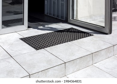threshold made of light gray ceramic tiles with steps at the entrance to the store with a foot mat and an open glass door at the facade of an office building close-up, nobody.