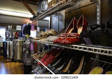 Thrift Store Racks Of Shoes With Clothing Racks In The Background.  Focal Point Is On Shiny Red Heels