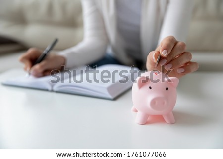 Thrifty woman sit at table hold pen writing daily expenses in diary put coin in pink piggy bank close up. Saving money for future, caring for tomorrow, makes stash of cash, investment, economy concept