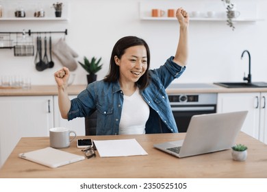 Thrilled asian woman raising hands in winner's gesture while looking at digital screen of computer in apartment. Emotional worker rejoicing over excellent news related to business partnerships.