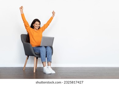 Thrilled emotional beautiful millennial hindu woman wearing casual outfit sitting in armchair over white blank wall, looking at computer laptop screen, gesturing raising hands up, copy space