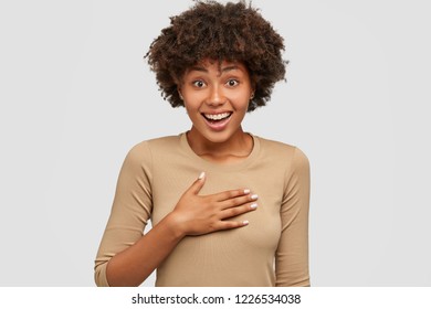 Thrilled Glad Dark Skinned Woman Keeps Hand On Chest, Surprised To Recieve Compliment From Stranger, Poses Against White Background, Has Toothy Tender Smile. People, Emotions And Feelings Concept.