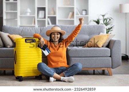 Thrilled happy beautiful young indian woman wearing comfy casual outfit and wicker hat sitting on floor with suitcase, holding passport and tickets, rasising hand up, home interior, copy space