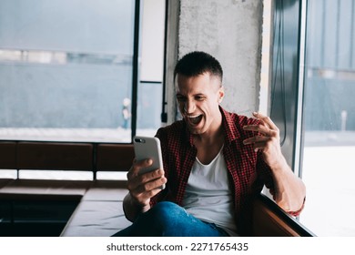 Thrilled male influencer receiving great news on internet while sitting near window using cellphone feeling euphoria after successful online work. Celebrating  sport betting win