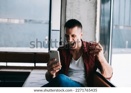 Thrilled male influencer receiving great news on internet while sitting near window using cellphone feeling euphoria after successful online work. Celebrating  sport betting win