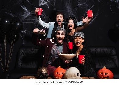 thrilled multicultural friends in halloween costumes near popcorn, carved pumpkins and skulls on black - Shutterstock ID 2044366970