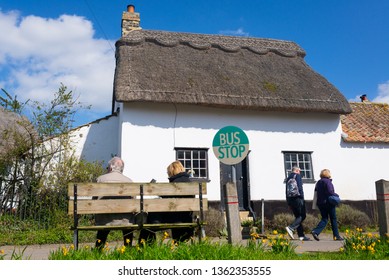 Thriplow, Cambridge, England, UK - March 2019: Senior Old Couple Sitting On A Wooden Bench At A Village Bus Stop With Traditional British English Cottage House And People Walking