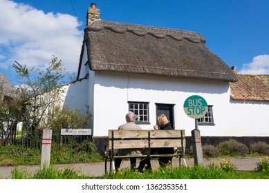 Thriplow, Cambridge, England, UK - March 2019: Senior Old Couple Sitting On A Wooden Bench At A Village Bus Stop With Traditional British English Cottage House With Thatched Roof