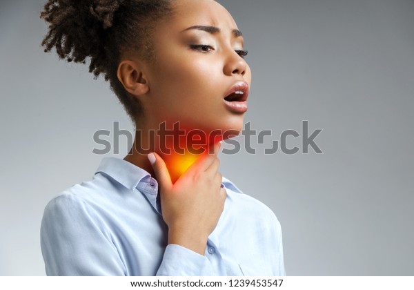Throat pain. Woman holding her inflamed throat.\
Photo of african american woman in blue shirt on gray background.\
Medical concept